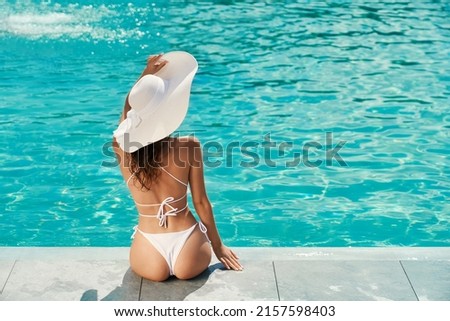 Back view of slim young female sitting near swimming pool. Pretty seductive female wearing white bikini and hat, looking forward, enjoying, relaxing. Concept of vacation.