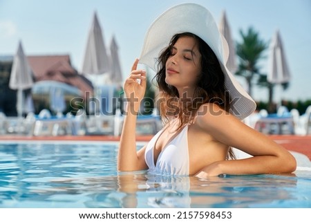 Front view of pretty brunette girl having vacation in nature. Seductive young female standing in water in pool, touching white hat, smiling with closed eyes. Concept of summer.