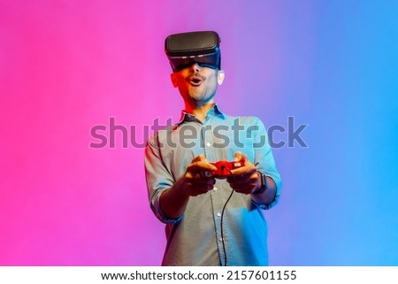 Portrait of amazed man in shirt in VR glasses playing video game, using simulator, yelling emotionally, being excited of gaming. Indoor studio shot isolated on colorful neon light background.
