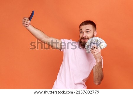 Portrait of positive bearded man making selfie photo with bunch of dollars on smartphone, bragging with wealth, wearing pink T-shirt. Indoor studio shot isolated on orange background. Royalty-Free Stock Photo #2157602779