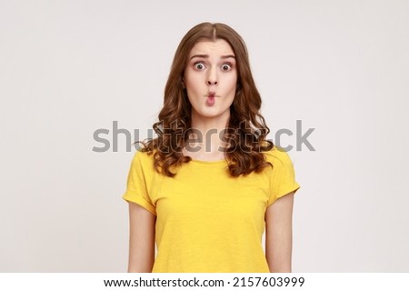 Portrait of charming funny teen girl with wavy hair in yellow T-shirt shows fish face grimace with pout lips, makes ridiculous childish comical grimace. Indoor studio shot isolated on gray background.