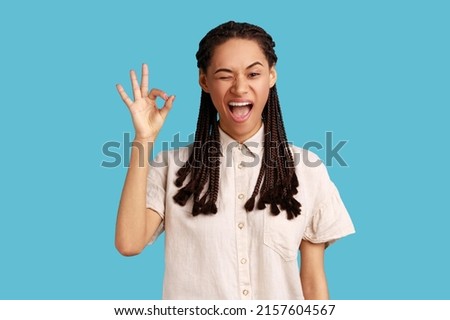 Portrait of cheerful young adult woman with black dreadlocks showing okay sign, assures you everything is fine, looks gladly, wearing white shirt. Indoor studio shot isolated on blue background.