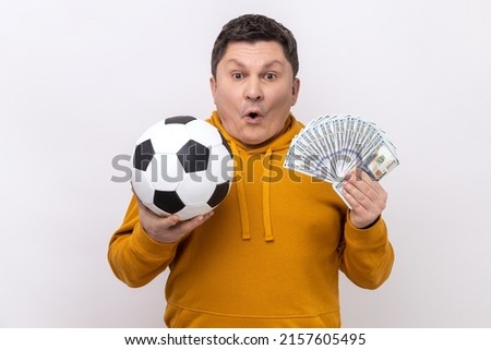 Surprised middle aged man showing soccer ball and fun of hundred dollar bills, winning lot of money betting for sport, wearing urban style hoodie. Indoor studio shot isolated on white background.