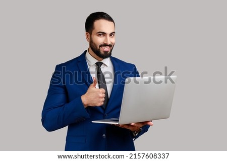 Portrait of optimistic bearded man holding laptop, looking at camera and showing thumb up, surfing internet, wearing official style suit. Indoor studio shot isolated on gray background.
