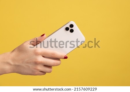 Closeup profile portrait of woman hand holding mobile phone, showing back side of cell phone. Indoor studio shot isolated on yellow background. Royalty-Free Stock Photo #2157609229