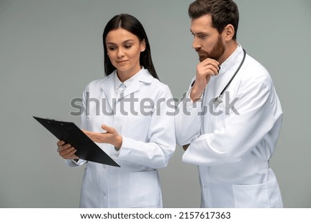 Female doctor holding clipboard and discussing something with her male colleague with stethoscope with concentrated faces while standing isolated on grey background 