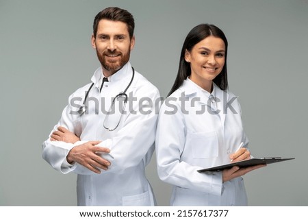 Woman doctor holding clipboard and posing with her male colleague with stethoscope with happy faces while standing isolated on grey background  Royalty-Free Stock Photo #2157617377