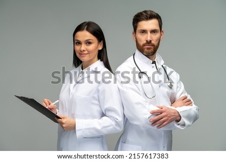 Woman doctor holding clipboard and posing with her male colleague with stethoscope with happy faces while standing isolated on grey background  Royalty-Free Stock Photo #2157617383