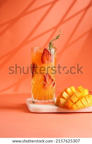 tall cocktail glass filled with yellow juice and fresh strawberries and cut in cubes mango half on tropical orange background, gobo mask effect