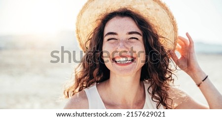 Young joyful woman in white shirt wearing hat smiling at camera on the beach - Traveler girl enjoying freedom outdoors on a sunny day - Wellbeing, healthy lifestyle and happy people concept