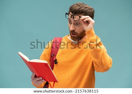 Shocking facts. Astonished smart bearded student man reading book with surprised expression, amazed by story, being shocked by interesting information. Indoor studio shot isolated on blue background  Royalty-Free Stock Photo #2157638115