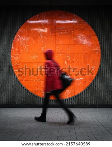 A vertical shot of a person walking near an orange circular mural in a station in blur motion