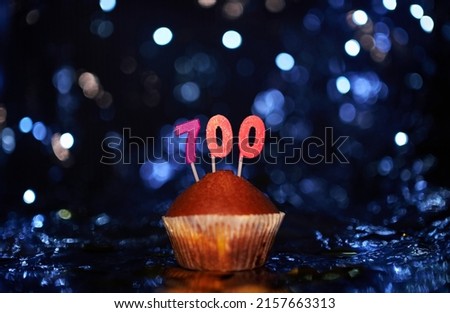 Digital gift card birthday concept. Tasty homemade vanilla anniversary cupcake with number 700 seven hundred on aluminium foil and blurred bright background in minimalistic style. High quality image