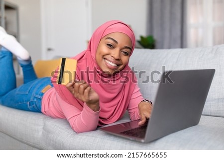 Easy payment and cashback concept. Happy african american lady doing online shopping at home using laptop and holding debit credit card in hand, smiling at camera while lying on sofa