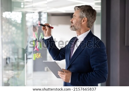 Business Planning. Mature Businessman Writing Ideas On Sticky Notes On Glass Wall At Office, Handsome Male Entrepreneur In Suit Holding Digital Tablet And Noting Information About Project, Copy Space Royalty-Free Stock Photo #2157665837