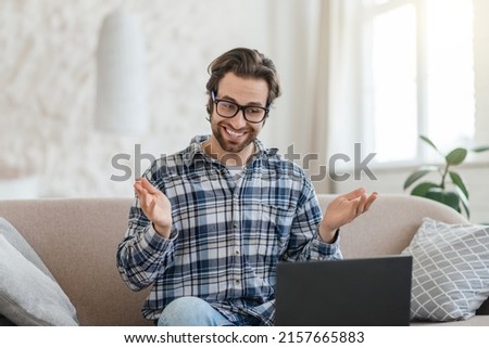 Cheerful young caucasian man teacher with stubble in glasses sits on sofa with laptop and gesticulates in living room interior. Study, education, video call, online lecture and work, covid-19 pandemic Royalty-Free Stock Photo #2157665883