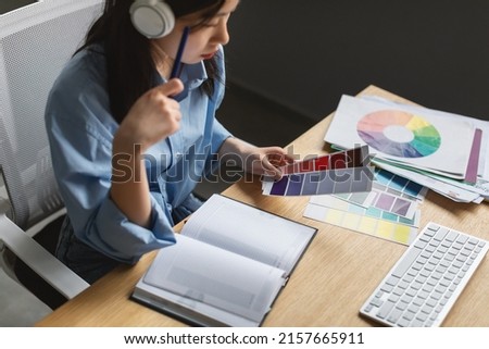 Autodidact Concept. Above top high angle view of busy focused Asian designer in wireless headset sitting at desk table with notebook and thinking, holding swatches choosing colors in modern office