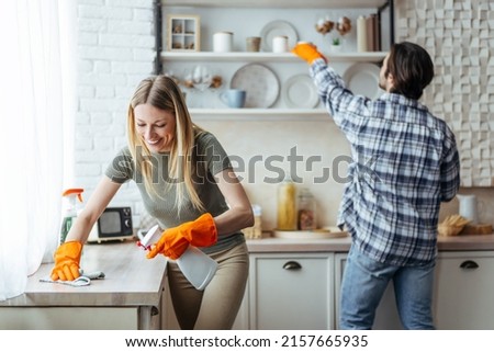 Smiling millennial european woman blonde with rubber gloves and man wipe dust on light kitchen interior, copy space. Hygiene, cleaning at home together and household chores during covid-19 outbreak Royalty-Free Stock Photo #2157665935