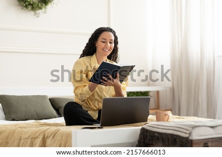 Smiling young woman sitting on bed at home, working on laptop taking notes in notebook, happy millennial female studying online, watching webinar using pc computer and writing check list