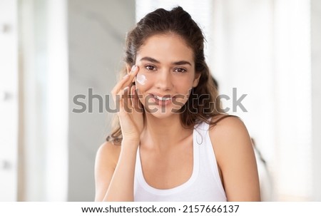 Portrait Of Attractive Woman Applying Facial Cream On Cheek Smiling To Camera Posing With Moisturizer On Face Standing In Modern Bathroom Indoor. Skincare Cosmetic Products Concept