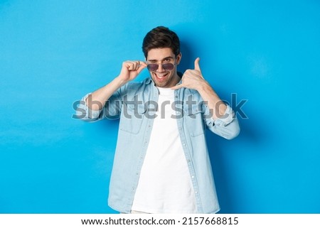 Handsome macho man in sunglasses flirting with you, make phone sign and winking, call me gesture, standing over blue background