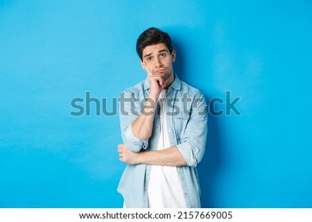 Unamused and bored man looking without interest at camera, standing against bllue background