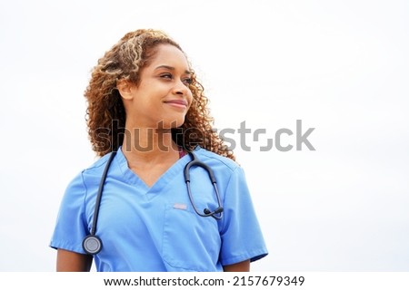 Beautiful young African American nurse or doctor with stethoscope stands, looking to the right side wearing blue scrubs and long curly hair                               