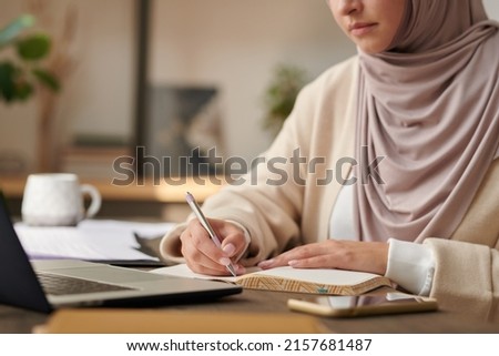 Unrecognizable Middle Eastern woman wearing headscarf working at office table writing something in her notebook
