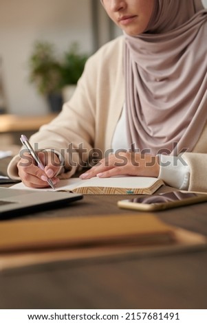 Middle Eastern woman wearing headscarf and abaya sitting at office table writing notes in her notebook, vertical head cropped shot