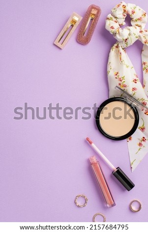 Makeup concept. Top view vertical photo of stylish scarf scrunchy lip gloss compact powder gold rings and two hairpins on isolated lilac background