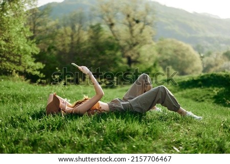 Happy woman blogger lying on the grass in the park and smiling with her phone in her hands against the backdrop of a summer natural landscape with sunlight Royalty-Free Stock Photo #2157706467