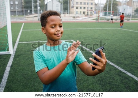 Kid using phone. Smiling and excited boy showing smartphone empty screen. Looking to camera. Cellphone display mockup mobile app advertisement. African american kid in football field. Blank screen
