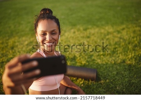 Smiling young African woman in sportswear sitting in a park listening to music on earphones and taking selfies after yoga practice