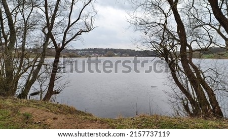 There are trees growing on the grassy shore of the lake. On the opposite shore is a village. It is overcast. It's raining and the water is covered in circles with its droplets Royalty-Free Stock Photo #2157737119