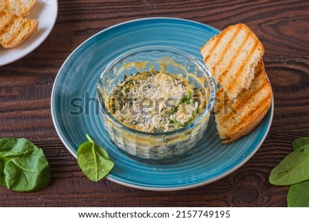 Hot appetizer, spinach and cream cheese dip in a transparent form on a blue plate on a brown wooden background. Served with toasted wheat bread. American food