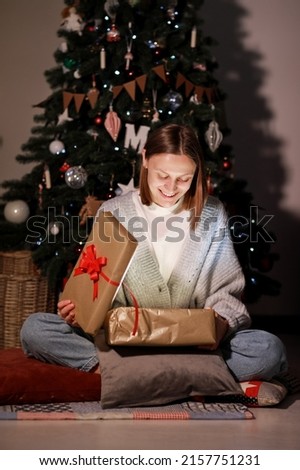 The Magic Of Christmas. Amazed young beautiful woman opening xmas gift box with light inside, sitting on floor near Christmas tree at home. High quality photo.