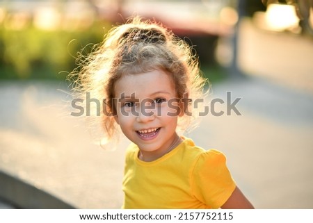 A little cute girl in a yellow T-shirt against the background of green trees.