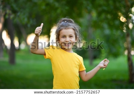 A little cute girl in a yellow T-shirt against the background of green trees.