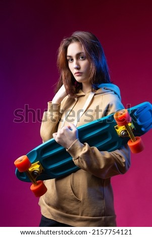 Young beautiful fashionable hipster girl dressed in a hoodie holds a skateboard in her hands on a purple studio background.