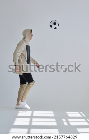 Young sportsman in street style wear training with soccer ball in large bright white hall with window shadow on the floor