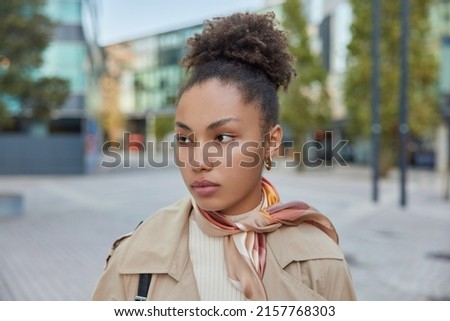 Thoughtful woman with curly combed hair wears coat and kerchief tied over neck concentrated away poses against blurred street background. Pretty female model explores new city during pastime
