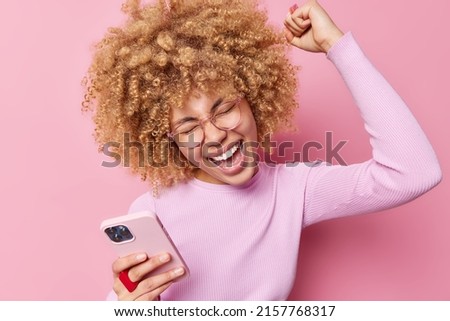 Beautiful curly haired young woman laughs positively shakes arm celebrates triumph uses modern smartphone cheers good news wears transparent eyeglasses casual jumper isolated over pink background. Royalty-Free Stock Photo #2157768317