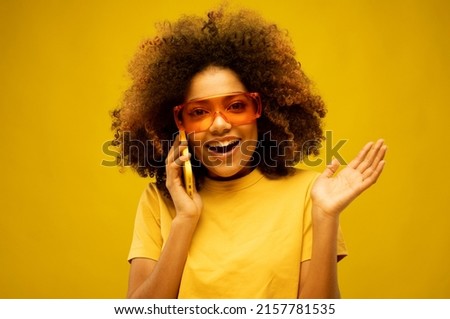 lifestyle, tehnology and people concept: Portrait of an excited young afro american woman wearing big sunglasses talking on mobile phone while standing isolated over yellow background