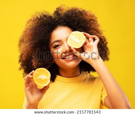 lifestyle, food, diet and people concept: Photo of young cheerful dark skin woman hold orange cover eye look empty space isolated on yellow color background