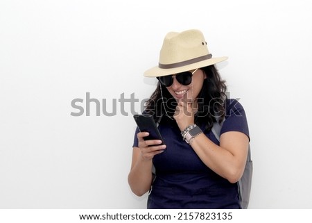Young latin woman with backpack, sunglasses and hat walks down the street like a tourist with a cell phone in her hand looking for information and taking photos
