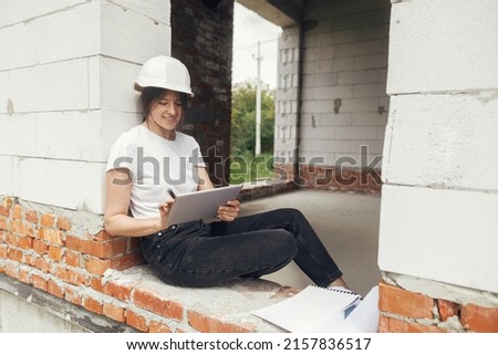 Young female architect with tablet checking blueprints while sitting in window of new modern house. Stylish woman engineer in hard hat looking at digital plans on tablet at construction site