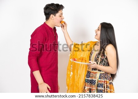 Young Indian brother and sister celebrating Diwali, rakshabandhan or bhai dooj festival isolated on white studio background. enjoying sweet laddo, Royalty-Free Stock Photo #2157838425