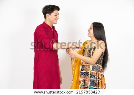 Happy indian teenage girl tying rakhi on young brother's hand occasion of rakshabandha or bhi dooj isolated on white studio background. both wearing traditional festive cloths. Royalty-Free Stock Photo #2157838429