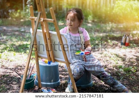 Little girl artist drawing a picture on canvas on an easel in nature, Cute little girl painting picture with a brush and color in backyard.  concept of art, hobby and relax