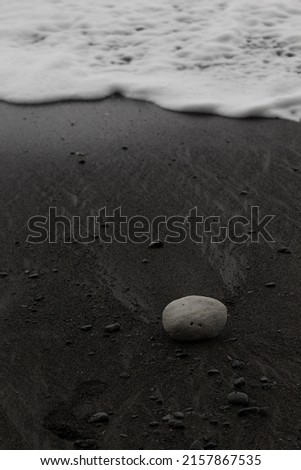 Wet black volcanic sand on seashore with white foam of wave as line and white stone, zen, texture, background, vertical.